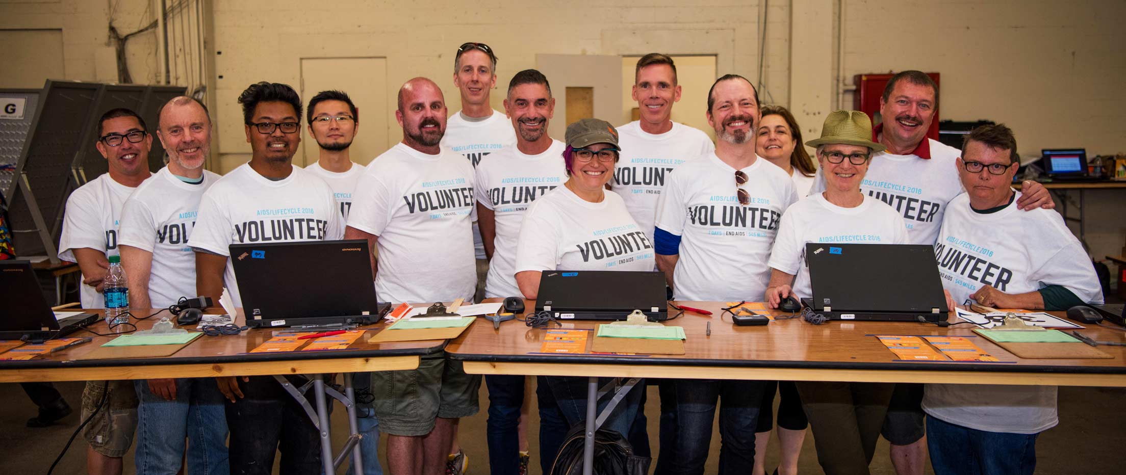 14 AIDS/LifeCycle Volunteers with smiling happy faces are ready to check you in at the Cow Palace. People are a diverse group wearing signature ALC Volunteer. You too can be a volunteer. See form link in navigation.