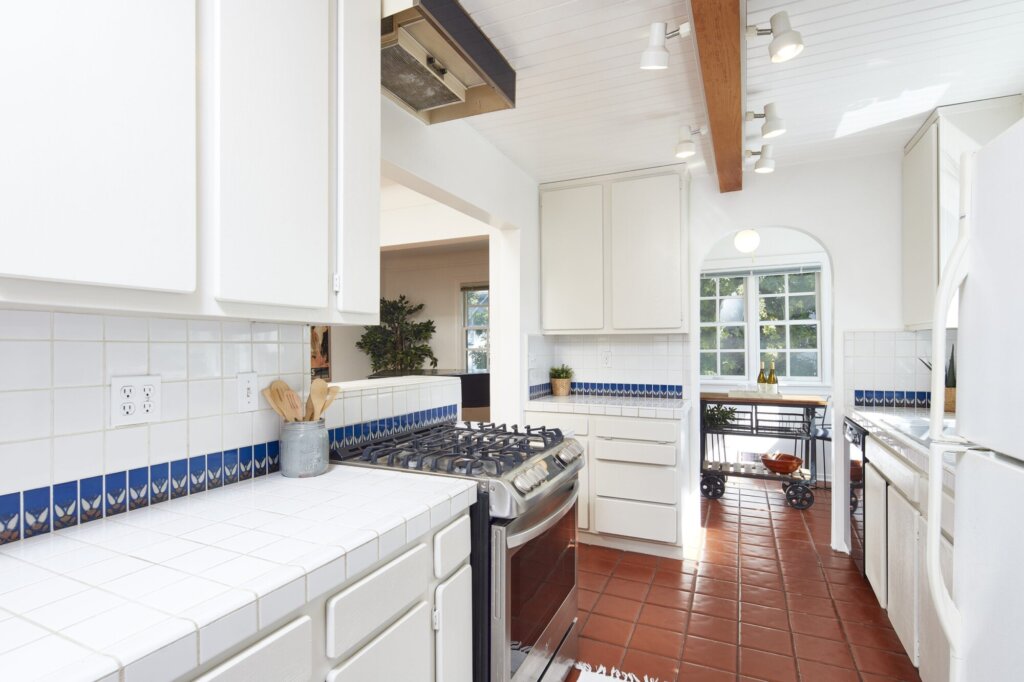 After: the same kitchen with white cabinets, a blue and white tiled backsplash and terracotta floors