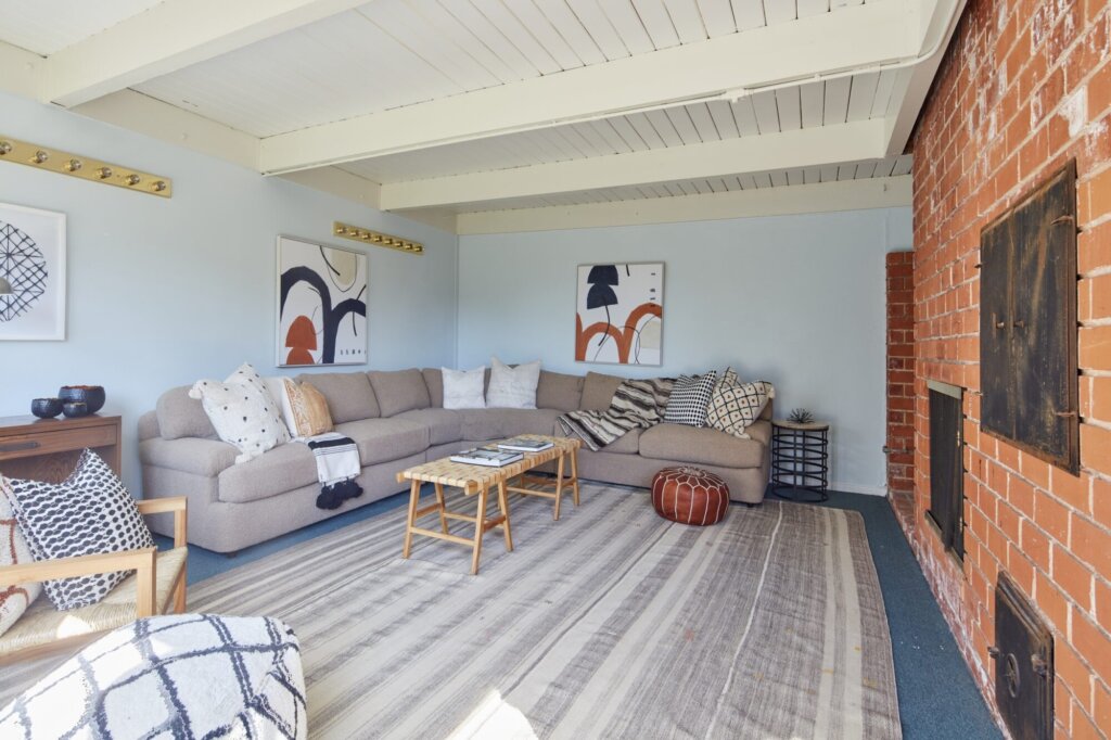 After: the same den with white beams, a sectional sofa and a styled brick fireplace